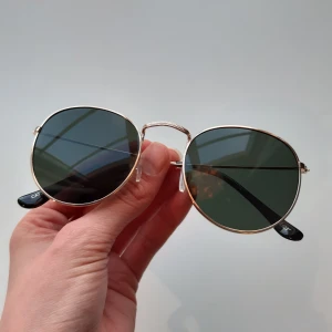 Solglasögon - Runda solglasögon med guldfärgad metallram och mörkgröna linser. De har justerbara näskuddar och svarta skalmar. Pris kan alltid diskuteras. Mycket bra skick!