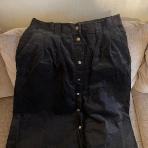 Svart manchesterkjol från Noa Noa - Snygg svart manchesterkjol från Noa Noa med knappar framtill. Kjolen har en klassisk design med en bekväm passform och är perfekt för kyligare dagar. Två fickor fram och en bak. 