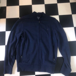 Blå tröja från Polo Ralph Lauren - Säljer en stilren blå tröja från Polo Ralph Lauren med dragkedja och broderad logotyp på bröstet. Tröjan har långa ärmar och är perfekt för en avslappnad look. Passar bra till både jeans och chinos.