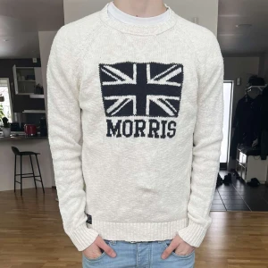 Vit stickad tröja från Morris - Säljer en stilren vit stickad tröja från Morris med ett stort Union Jack-motiv och texten 'Morris' på framsidan. Tröjan har långa ärmar och ribbade muddar vid ärmslut och nederkant. Perfekt för en klassisk look.