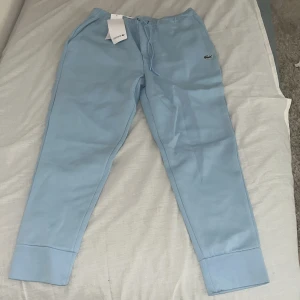 Ljusblå mjukisbyxor från Lacoste - Säljer ett par ljusblå mjukisbyxor från Lacoste med dragsko i midjan och den klassiska krokodilloggan på benet. Perfekta för en avslappnad stil.