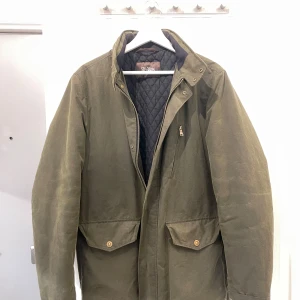 Grön jacka med dragkedja - Säljer en stilren grön jacka med dragkedja och knappar. Jackan har en quiltad insida och praktiska fickor framtill. Perfekt för kyligare dagar.