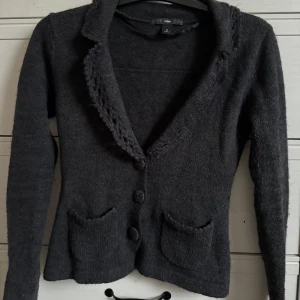 Mörkgrå stickad kofta från Hm med fickor och knappar - Säljer en snygg mörkgrå stickad kavaj från Vero Moda. Den har en elegant krage och knäpps med tre knappar framtill. Kavajen har två fickor och är tillverkad i en mjuk blandning av mohair och akryl. Perfekt för kyligare dagar!