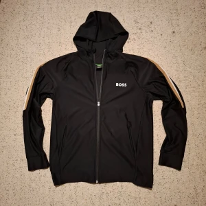 Svart huvtröja från BOSS - Snygg svart huvtröja från BOSS med dragkedja och coola ränder i beige och vit längs ärmarna. Perfekt för en sportig look. Huvan ger extra stil och funktionalitet. Pris kan diskuteras