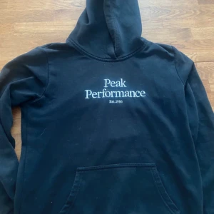 Svart hoodie från Peak Performance - Säljer en svart hoodie från Peak Performance med tryck på framsidan. Den har en klassisk design med huva och en stor ficka framtill. Perfekt för en avslappnad stil.