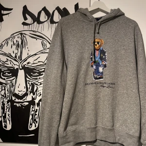Grå hoodie från Ralph Lauren - Säljer en grå hoodie från Ralph Lauren med en Polo Bear-design på framsidan. Tröjan har en klassisk huva och långa ärmar, perfekt för en avslappnad stil. Den är gjord i mjukt material för extra komfort. Perfekt för dig som vill ha en stilren och bekväm look. Nypris ligger på 1799kr och den är sparsamt använd 