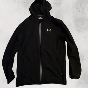 Under armour reflective windbreaker - Säljer en svart träningsjacka från Under Armour med dragkedja och huva. Jackan har långa ärmar och reflekterande detaljer längs dragkedjan. Perfekt för löpning och andra utomhusaktiviteter.