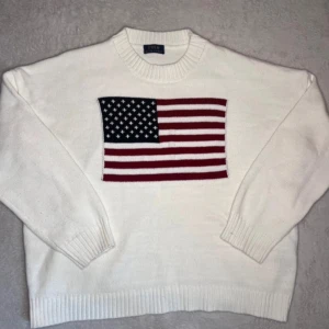 Vit stickad tröja från Polo Ralph Lauren - Vit stickad tröja från Polo Ralph Lauren med en stor amerikansk flagga på framsidan. Tröjan har långa ärmar och ribbstickade muddar vid ärmslut och nederkant. Perfekt för en stilren och patriotisk look.