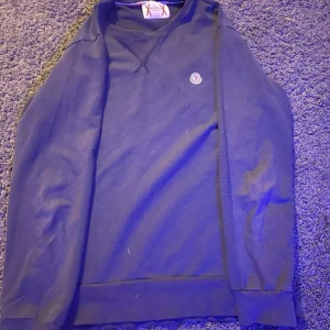 Moncler tröja - Säljer en snygg mörkblå tröja från Moncler med deras ikoniska logga på bröstet. Tröjan har en klassisk design med rund hals och långa ärmar. Perfekt för en stilren look.