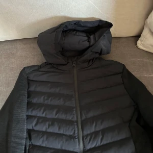 Svart jacka från Engelbert Strauss - Säljer en svart jacka från Engelbert Strauss med huva och dragkedja. Jackan har en pufferdesign på framsidan och ribbade ärmar. Perfekt för kyligare dagar!