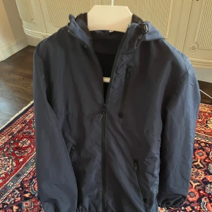 Mörkblå vindjacka från H&M Divided - Säljer en mörkblå vindjacka från H&M Divided. Jackan har en dragkedja framtill och en praktisk huva. Perfekt för blåsiga dagar och har en stilren design med långa ärmar.