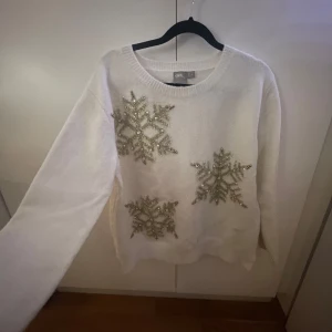 Vit tröja med snöflingor från ASOS - Säljer en vit tröja från ASOS med glittriga snöflingor och pärlor/stenar på framsidan. Tröjan har långa ärmar och är perfekt för vintersäsongen. Den är tillverkad i en mjuk och bekväm polyesterblandning.