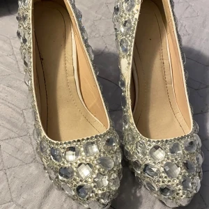 Glittriga klackar med stora stenar - Säljer ett par riktigt glittriga klacksr täckta med stora, klara stenar och pärldetaljer. Skorna har spetsig tå och hög klack, perfekta för att sticka ut på festen. Färgen är silver och materialet är syntet.