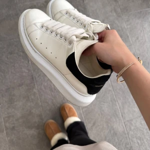 Vita sneakers från Alexander McQueen med svart häl - Säljer ett par klassiska vita sneakers från Alexander McQueen med svart mockadetalj på hälen och tjock vit sula. Skorna har snörning och diskreta hål på sidorna för ventilation. Perfekta för en stilren och trendig look.