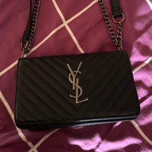 Svart axelväska - Säljer en svart quiltad axelväska från Ysl (inte äkta) med silverfärgad kedja och YSL-logga framtill. Väskan har stilrena linjer och klassisk design, perfekt för att lyfta vilken outfit som helst.