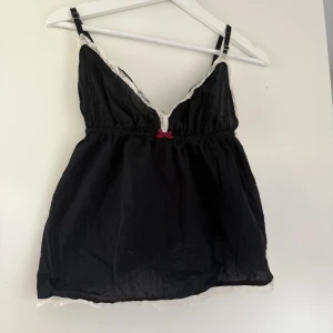 Svart linnetopp med spets från Odd Molly - Supersöt svart linnetopp från Odd Molly med vita spetsdetaljer längs kanterna och en liten rosa rosett framtill. Toppen har smala axelband och är tillverkad i ett tunt, mjukt material. Perfekt att styla lager på lager eller som den är. Använd enstaka gång 