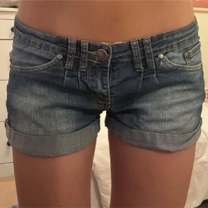 Blå jeansshorts med upprullade ben - Snygga blå jeansshorts från One Love Denim med upprullade ben och klassisk femficksdesign. Shortsen har låg midja och stängs med knapp och dragkedja. Perfekta för varma dagar och enkel att matcha med olika toppar.