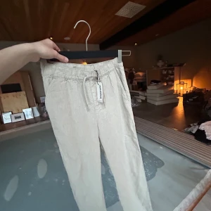 Beiga linnebyxor med dragsko från Kappahl - Snygga och luftiga beiga linnebyxor från Kappahl med elastisk midja och dragsko. Perfekta för varmare dagar och har en avslappnad passform. Byxorna har raka ben och är tillverkade i ett lätt material.