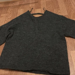 Mörkgrå stickad tröja - Mysig mörkgrå stickad tröja med öppen rygg och långa ärmar. Tröjan har en enkel och avslappnad passform, perfekt för kyligare dagar. Passar bra till jeans eller kjol för en stilren look.