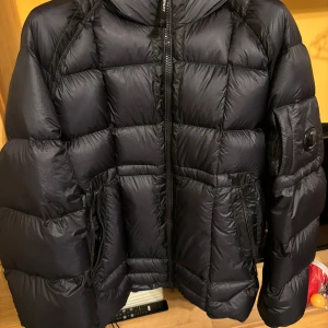 Svart pufferjacka från C.P. Company - Säljer en svart pufferjacka från C.P. Company i storlek L. Jackan har lång ärm, dragkedja framtill och två fickor med dragkedja. Klassisk badge på ärmen och quiltad design som ger en modern look.