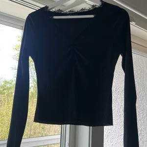 Svart långärmad topp med spetsdetalj - Svart långärmad topp med v-ringning och spetsdetalj längs halsringningen. Toppen har en lätt rynkning framtill och är figurnära i modellen. Perfekt att styla till olika outfits.