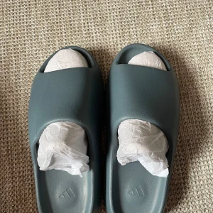 Adidas yeezy  slides i mörkgrått - Säljer ett par mörkgrå Adidas slides med öppen tå och räfflad sula. De är tillverkade i syntetmaterial och har en minimalistisk design med Adidas-logga på fotbädden. Perfekta för sommaren eller att chilla hemma. Dm för frågor👌😀