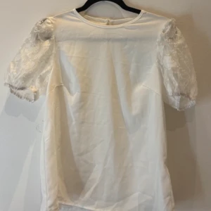 Vit blus med spetsärmar - Säljer en söt vit blus med puffiga spetsärmar och rund halsringning. Blusen har en liten knapp i nacken och är perfekt för dig som gillar romantiska detaljer. Ärmarna är korta och dekorerade med vacker spets.