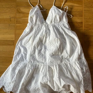 Vit somrig klänning från Zara med broderade detaljer - Säljer en vit klänning från Zara med tunna axelband och v-ringning. Klänningen har broderade spetsdetaljer och volang nertill, perfekt för varma dagar. Lätt och luftig modell med omlottkänsla och romantisk look.