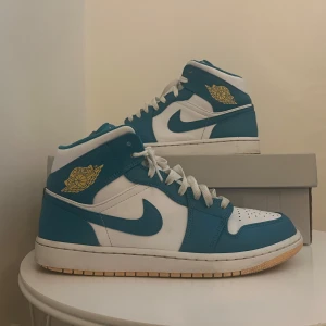 Nike Air Jordan 1 Mid Baby Blue - Säljer ett par Nike Air Jordan 1 Mid i turkos och vitt med klassisk Air Jordan-logga i gult på sidan. Skorna har hög modell, snörning och vit sula med turkosa detaljer. Perfekta för dig som gillar streetwear och letar efter unik sommar skor