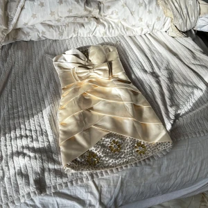 Beige kort klänning med rosett och pärldetaljer - Säljer en champagnefärgad kort klänning i satin med vacker rosett framtill. Klänningen har axelbandslös design, lager på lager-drapering och nedtill finns pärlor och blommiga dekorationer i gult och silver. Perfekt för speciella tillfällen!