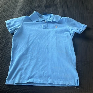 Ljusblå pikétröja från GANT - Säljer en klassisk ljusblå pikétröja från GANT i bomull. Tröjan har korta ärmar, krage och knappar framtill samt en liten broderad logga på bröstet. Perfekt för en stilren och avslappnad look.