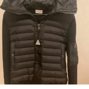 Svart pufferjacka från Moncler - Säljer en svart pufferjacka från Moncler med stickade ärmar och huva. Jackan har dragkedja framtill, två fickor med knappar och en ärmficka med dragkedja. Perfekt för kyliga dagar och snygg till vardags.