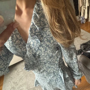 Ljusblå blommig omlottklänning med volang - Supersöt ljusblå kortklänning med vitt blommönster och volangdetaljer. Omlottmodell med v-ringning och långa ärmar. Perfekt för vår och sommar, ger en romantisk och fräsch look.💕