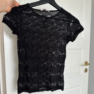 Svart spetsblus - Säljer en svart spetsblus med korta ärmar och små knappar framtill. Blusen har ett feminint och transparent utseende med fina spetsdetaljer över hela plagget. Perfekt nu till sommaren. Skulle säga storlek xs/s.