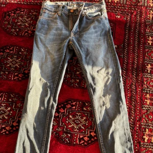 Blå jeans från Nudie Jeans - Säljer ett par klassiska blå jeans från Nudie Jeans med raka ben och snygga kontrastsömmar. Byxorna har fem fickor, gylf med dragkedja och Nudie-logga på bakfickan. Perfekta för en stilren vardagslook. Aldrig använda storlek w30/l32