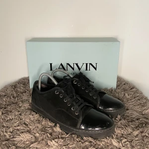 Lanvin Cap Toe - Säljer ett par svarta sneakers från Lanvin med snygg lackad tå och snörning. Skorna har en klassisk rund tå och platt sula, perfekt för en stilren look. Kommer med originalkartong.
