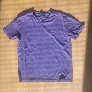 Lila ralph lauren T-Shirt -  Ralph Lauren t-shirt i snygg lila färg med den klassiska gula broderade polospelaren på bröstet, storlek M. En ikonisk t-shirt med hög kvalitet och tidlös design – perfekt inför sommaren! Tyget är mjukt, andas bra och sitter skönt, oavsett om du ska ut och resa, gå på stan eller bara njuta av solen. Den är använd men i mycket bra skick, utan fläckar eller hål. Nypris ligger runt 899 kr, så mitt pris på 2