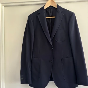 Mörkblå ullkavaj från Z Zegna - Snygg mörkblå kavaj från Z Zegna i 100% ull. Klassisk modell med två knappar framtill, tre fickor och slits bak. Perfekt för dig som vill ha en stilren och tidlös look. Nypriset är väldigt högt…