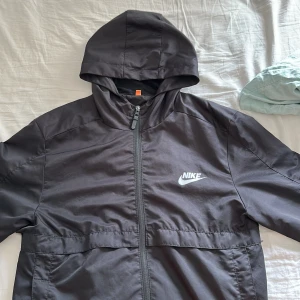 Svart vindjacka med huva från Nike - Säljer en svart vindjacka från Nike med huva och dragkedja framtill. Jackan har två sidofickor och klassisk Nike-logga på bröstet. Perfekt för blåsiga dagar och enkel att matcha med det mesta.