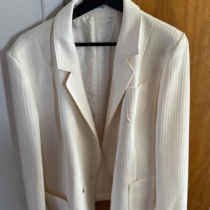 Vit kavaj med diskreta ränder - Säljer en stilren vit kavaj med subtilt randigt mönster. Klassisk krage, tre fickor framtill och snygg passform. Perfekt för dig som vill ha en fräsch och clean look.