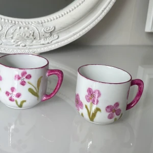 Handmålade espressokoppar med blommor - Två små handmålade espressokoppar i porslin med rosa blommor och gröna blad. Kopparna har rosa handtag och kant, signerade Brit 1999 under botten. Perfekta för samlare eller som dekoration.