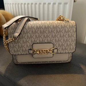 Beige axelväska från Michael Kors - Snygg beige axelväska från Michael Kors med MK-logga över hela väskan och guldfärgade kedjedetaljer. Väskan har ett dekorativt spänne framtill med MK-emblem och justerbar axelrem. Perfekt för att lyfta din outfit!