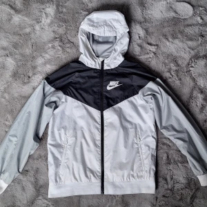 Nike vindjacka med huva stl 147-158  - Snygg vindjacka från Nike i svart, grått och vitt med dragkedja och huva. Jackan har två fickor med dragkedja och klassisk Nike-logga på bröstet. Perfekt för dig som vill ha en stilren och sportig look.