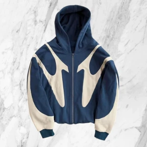 Blå hoodie med vita detaljer och dragkedja - Säljer en blå hoodie med unika vita mönster och dragkedja framtill. Jackan har huva, långa ärmar och ribbade muddar. På ryggen finns ett vitt kors som detalj. Perfekt för dig som vill sticka ut med din stil.