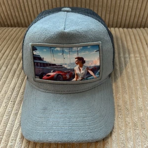 Grå keps med retro bilmotiv - Snygg grå keps med mesh baktill och ett stort tygmärke framtill med ett retroinspirerat motiv av en kvinna och en röd sportbil.