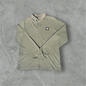 Olivgrön pikétröja från Stone Island - Säljer en olivgrön pikétröja från Stone Island med klassisk krage och knäppning framtill. Tröjan har lång ärm och en svart logotyp-patch på bröstet. Perfekt för en stilren och avslappnad look. 