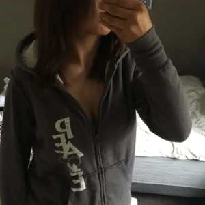 Mörkgrå hoodie från Cocoa Platinum - Mysig mörkgrå hoodie med vit teddyfoder i luvan och insidan. Dragkedja framtill och två fickor. Tryck i vitt längs ena sidan framtill. Perfekt för kyliga dagar och avslappnad stil.