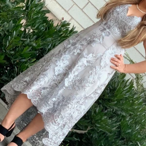 Silverfärgad spetsklänning med blommigt mönster - Säljer en elegant silverfärgad klänning med vackert blommigt spetsmönster och v-ringad halsringning. Klänningen har ärmlös design och en lätt utsvängd kjol som ger en lyxig känsla. Perfekt för speciella tillfällen!