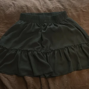 Svart volangkjol från Sister - Supersöt svart kortkjol från Sister med bred resår i midjan och volangdetalj runt om. Kjolen har en luftig och flowy känsla, perfekt för att styla med en enkel topp eller hoodie.