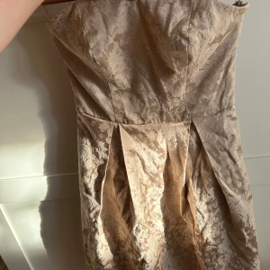 Champagnefärgad axelbandslös klänning från Vila - Säljer en elegant axelbandslös klänning från Vila i champagnefärgat tyg med vackert blommigt mönster. Klänningen har veckad kjol och smock baktill för extra komfort. Perfekt för speciella tillfällen!